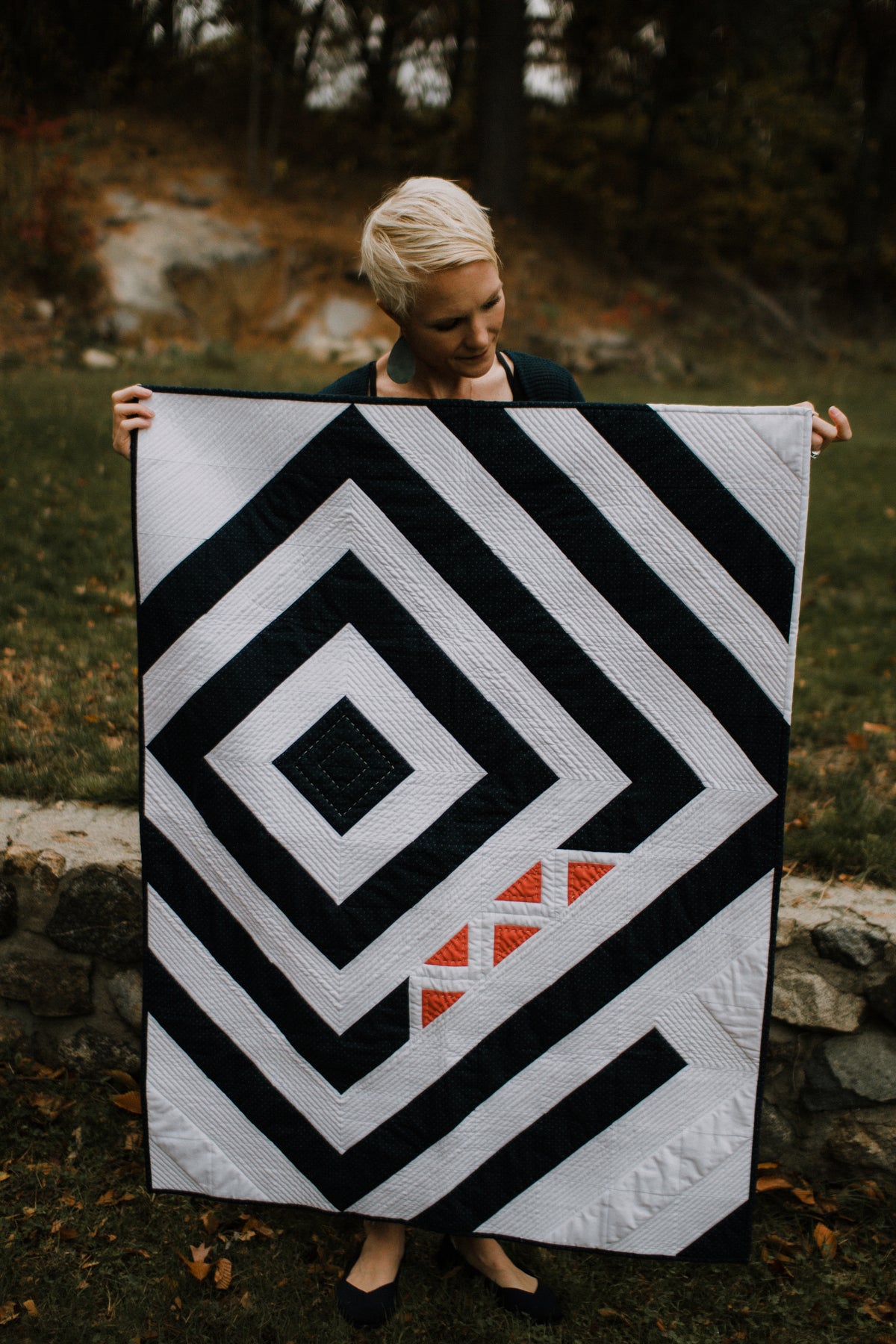 A woman with short blonde hair holds up a large quilted blanket outdoors. The blanket features a bold black and white geometric pattern of nested diamonds and concentric squares, with a small orange accent of three triangles near the bottom right.